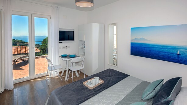 A bedroom with a bed, a desk, and a TV. There is a balcony with a view of the sea and mountains. - Maison Cipriani (Massa Lubrense)