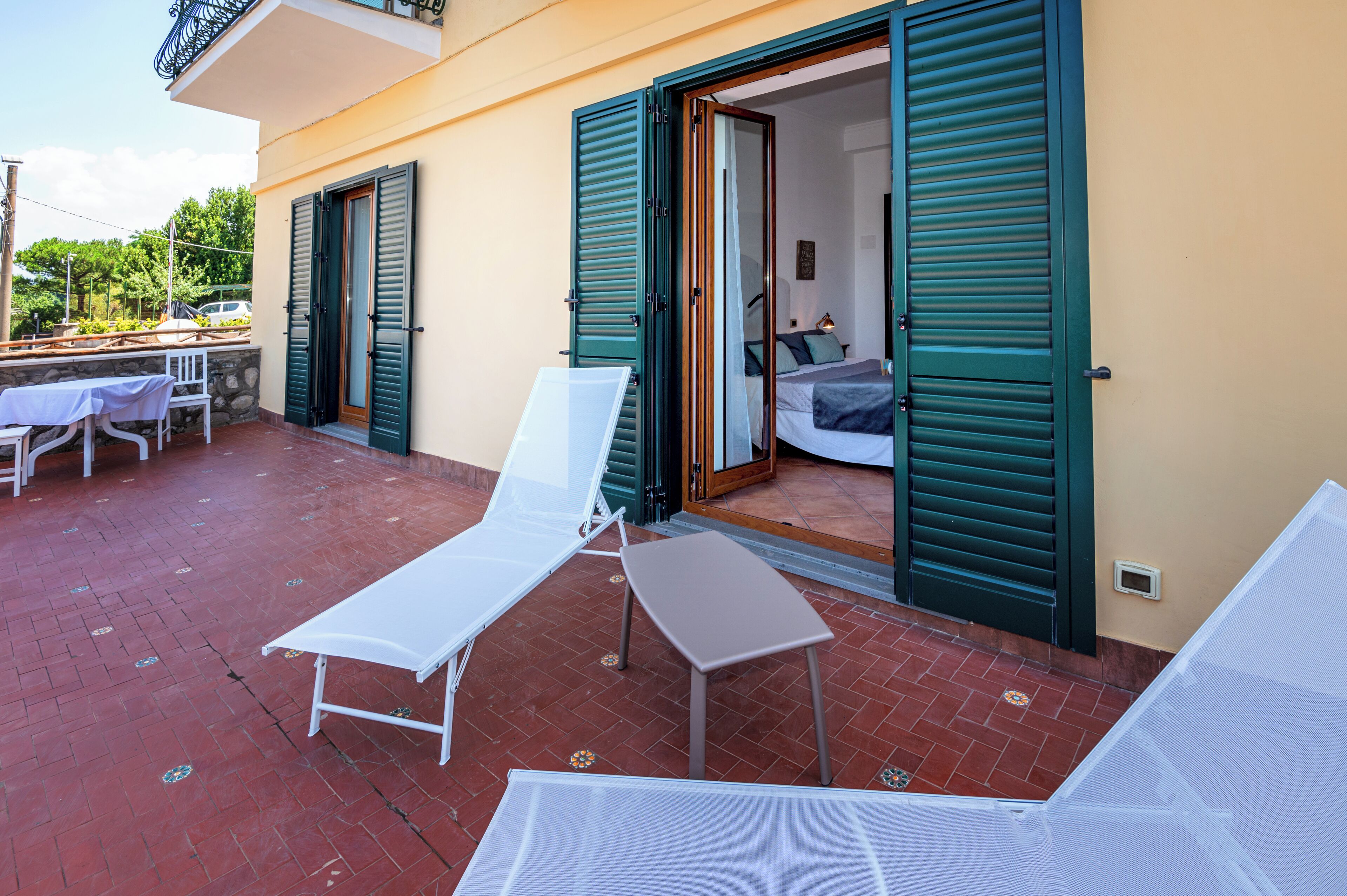 A balcony with two white lounge chairs, a small table, and a partially open door leading to a bedroom with a bed and a nightstand.
