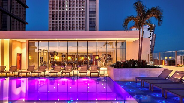 Un hôtel moderne doté d’une piscine sur le toit, agrémentée d’éclairages bleus et violets, de chaises longues et d’un palmier.