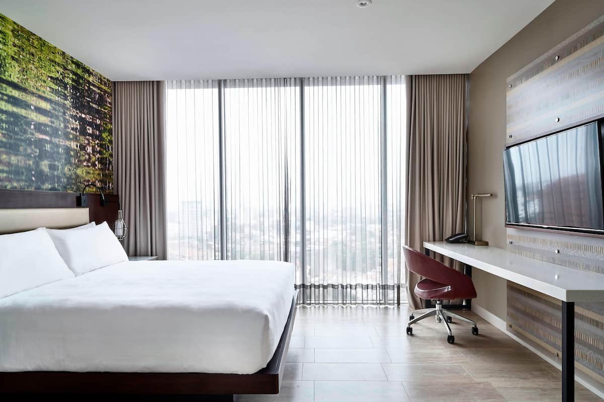 Un hotel moderno con una cama grande, un escritorio, un televisor y vistas a la ciudad a través de ventanales.