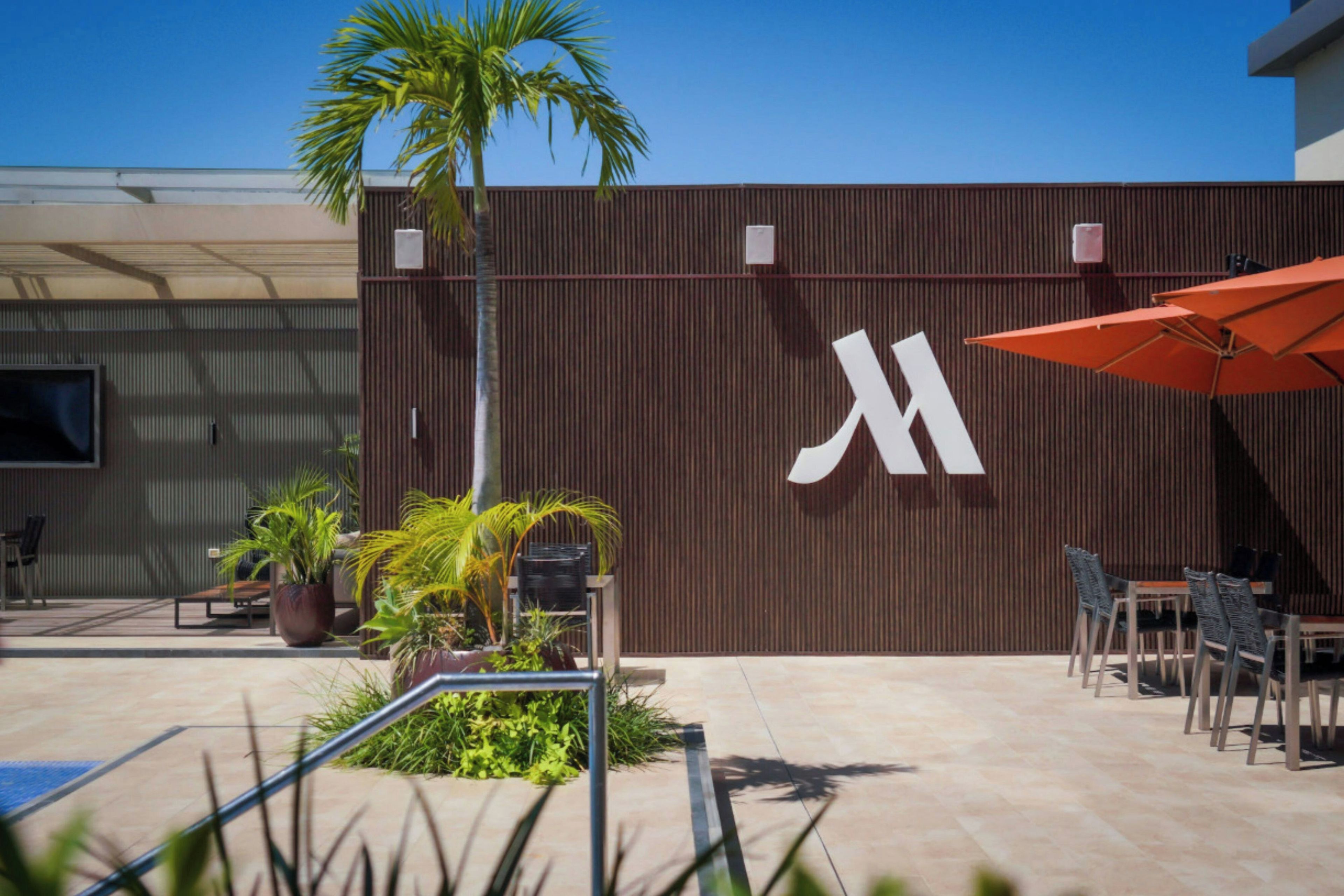 Un espace extérieur moderne, aménagé avec des sièges, une piscine, des palmiers et un grand bâtiment orné d’un logo « M » bien visible.