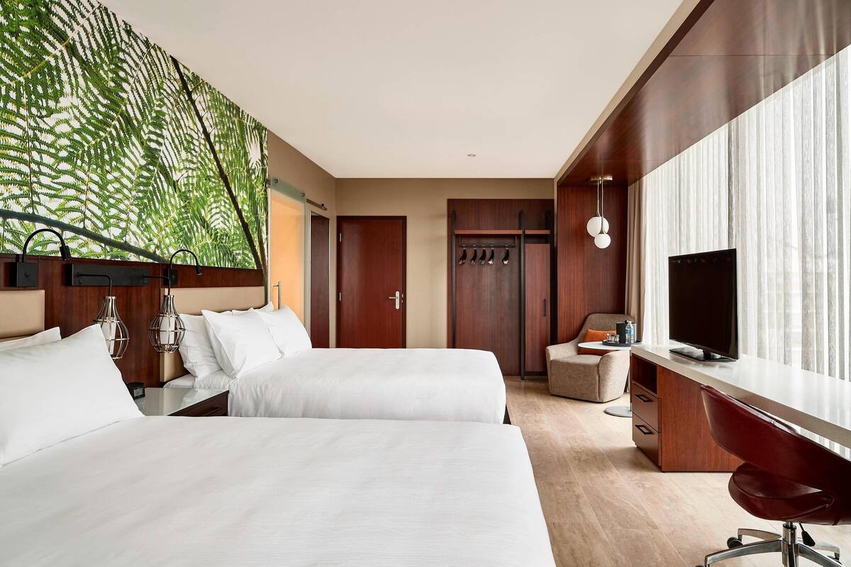 Habitación de hotel con dos camas, un escritorio, un televisor y un ventanal grande con cortinas.
