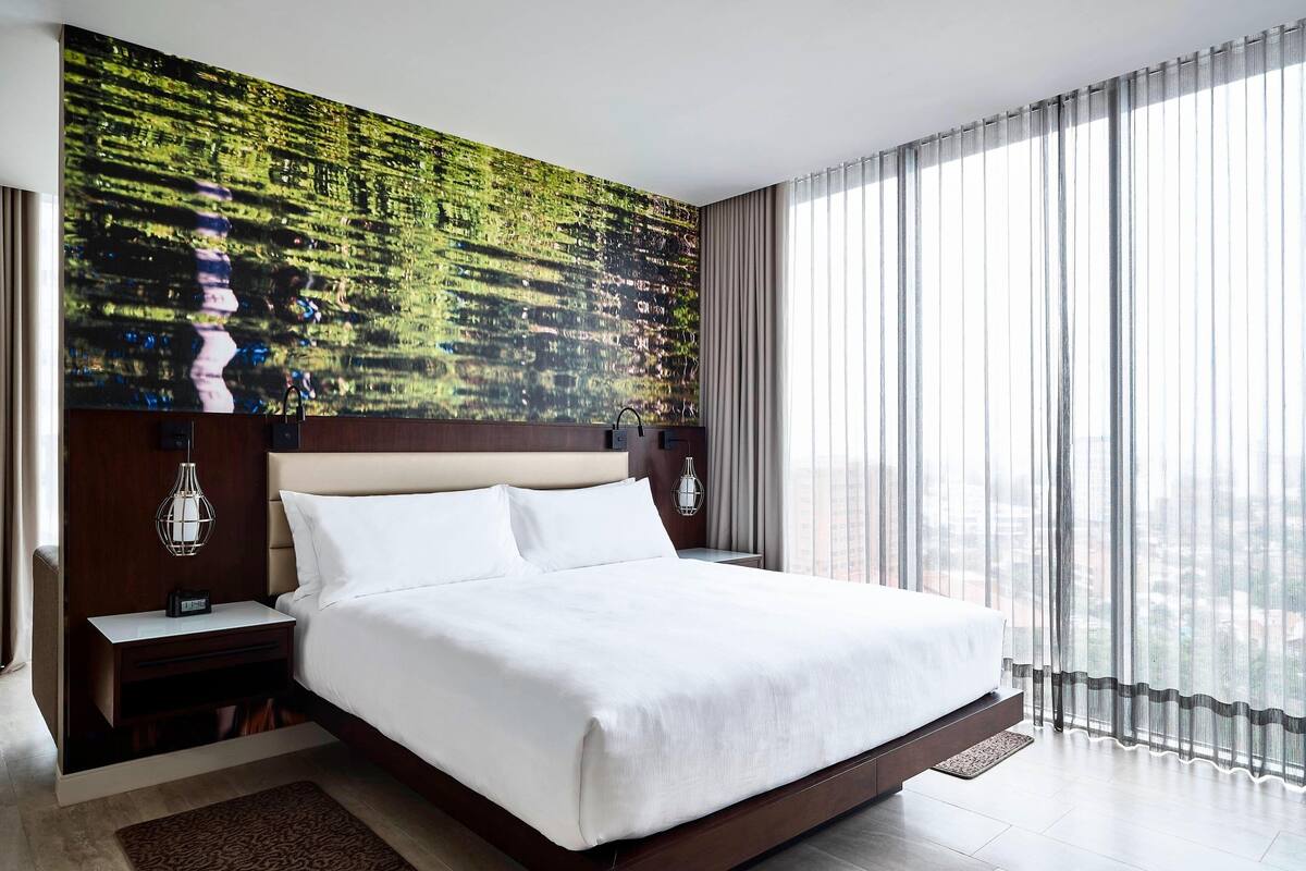 Habitación de hotel moderna con una cama grande, una mesita de noche con lámpara, vistas de la ciudad a través de cortinas traslúcidas y un panel decorativo en la pared.