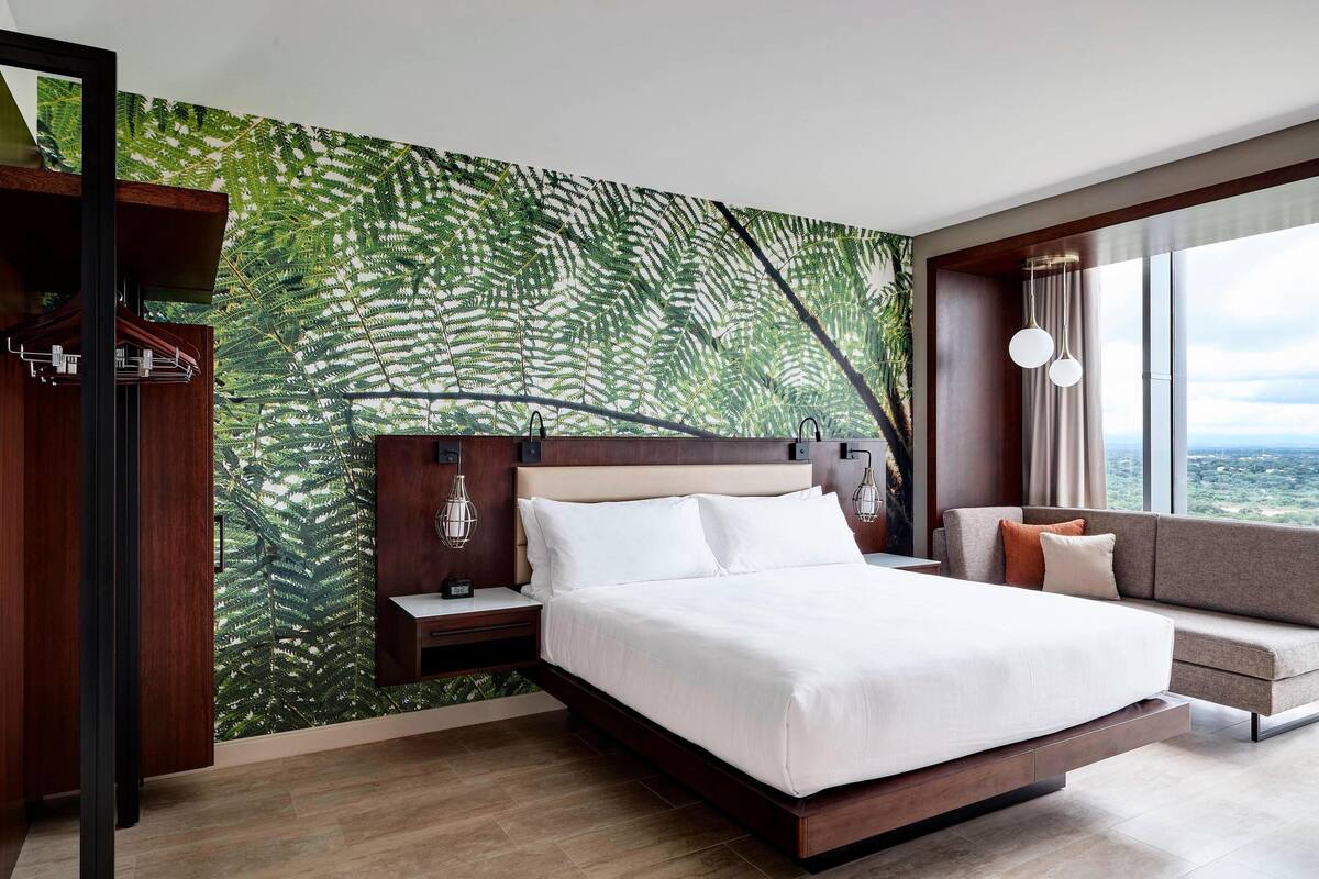 Habitación de hotel moderna con una cama grande, un sofá, una mesita de noche y una pared con estampado de hojas de helecho.