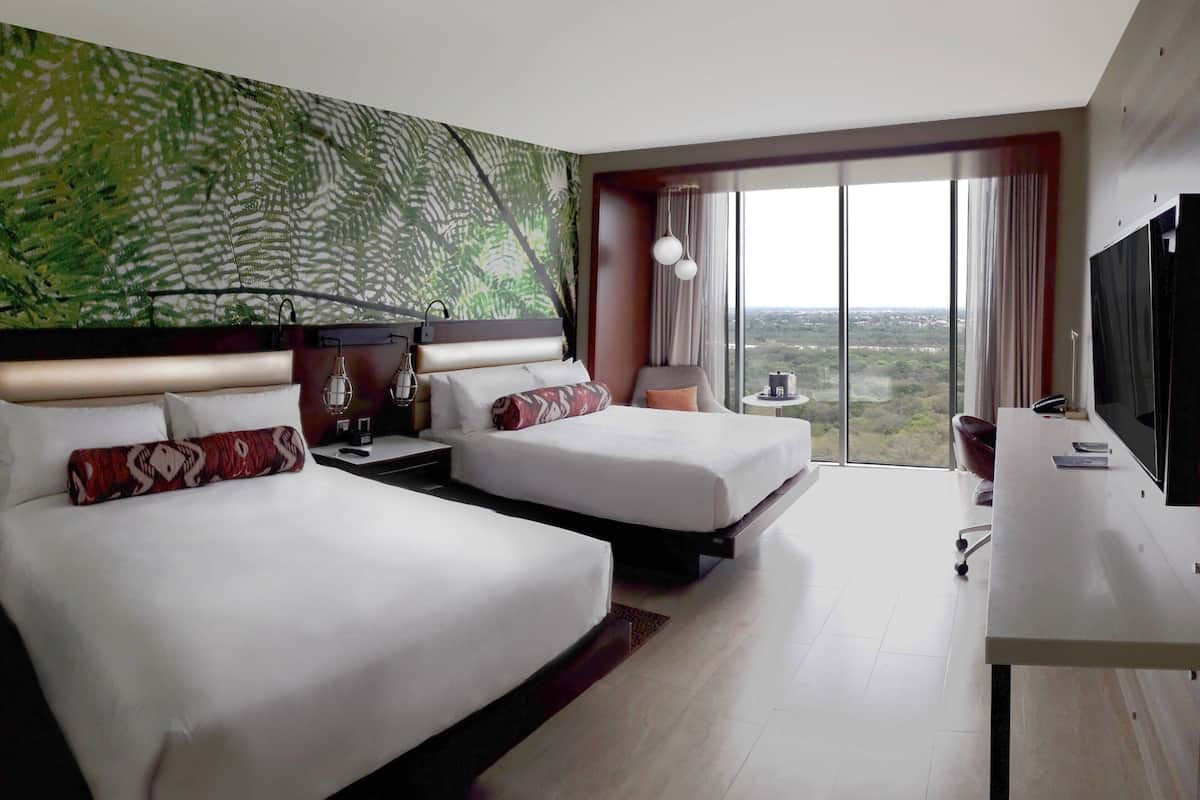 Habitación de hotel con dos camas, un escritorio, un televisor y un ventanal con vistas a la ciudad.