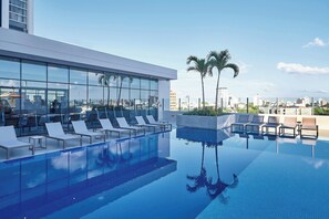 Piscina en la azotea con sillones de descanso y palmeras, rodeada de edificios modernos.
