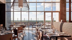 Un restaurant avec un plafond haut, de grandes fenêtres et une vue sur le paysage urbain. Les tables sont dressées avec des assiettes, des verres et des couverts.