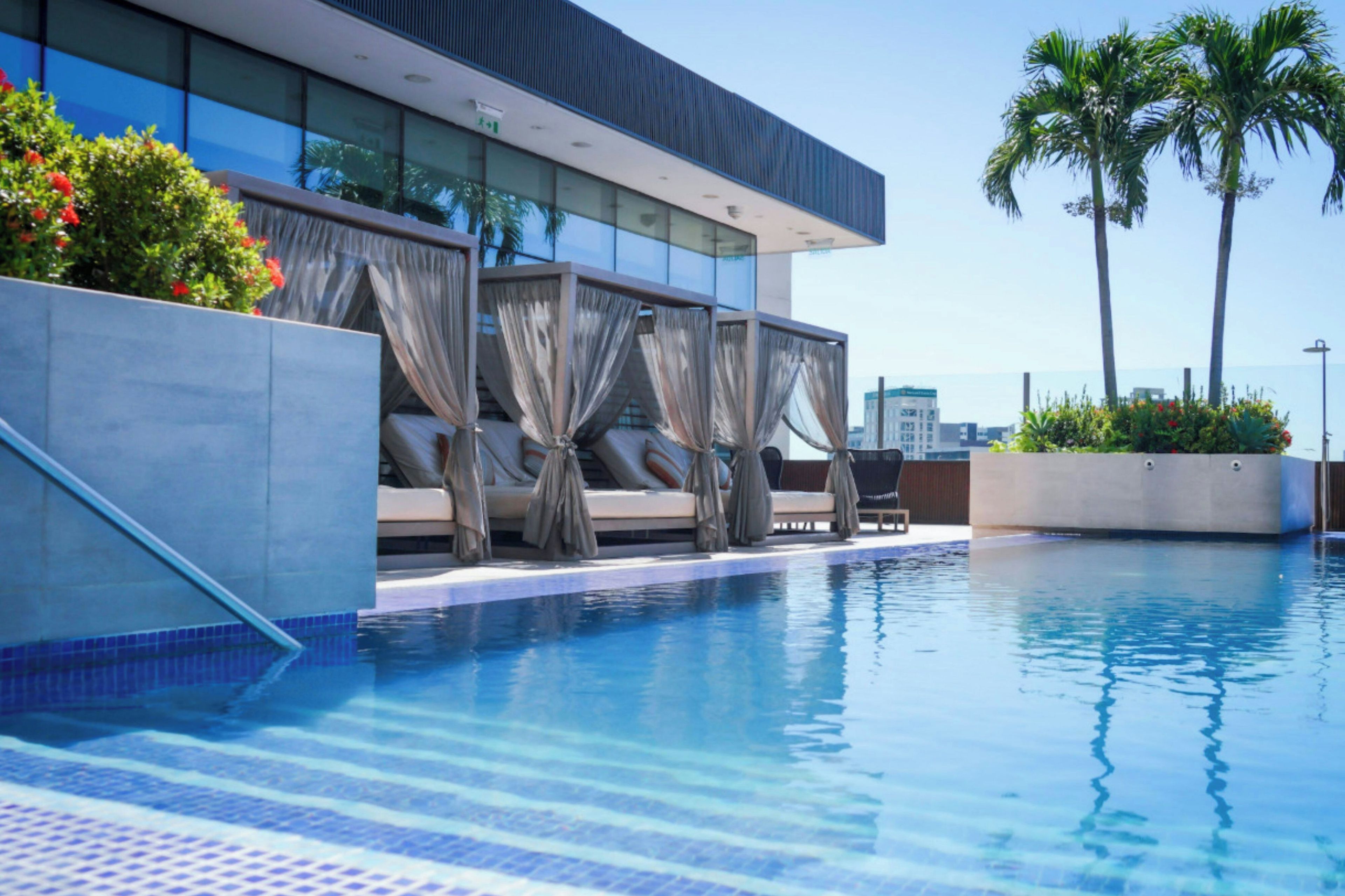 A pool area with a modern building, a seating area with umbrellas, and palm trees.