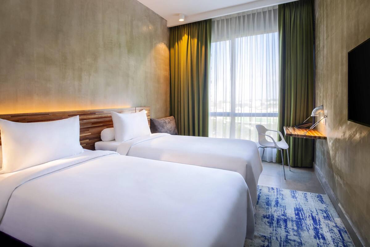 A hotel room with two beds, a desk with a chair, a TV, and a large window with curtains.