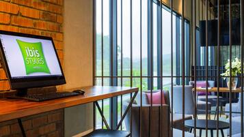 A hotel reception desk with a computer displaying 'ibis Styles Hotels' logo, a brick wall, large windows, and a seating area with tables and chairs.