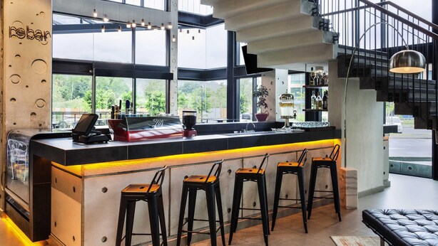 A modern bar with a long counter, bar stools, and a staircase leading to an upper level. - Ibis Styles Kota Kinabalu Inanam Hotel (Kota Kinabalu)