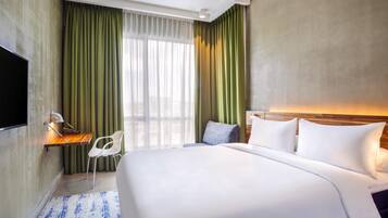 A hotel room with a large bed, a desk with a chair, a flat-screen TV mounted on the wall, and a window with green curtains.