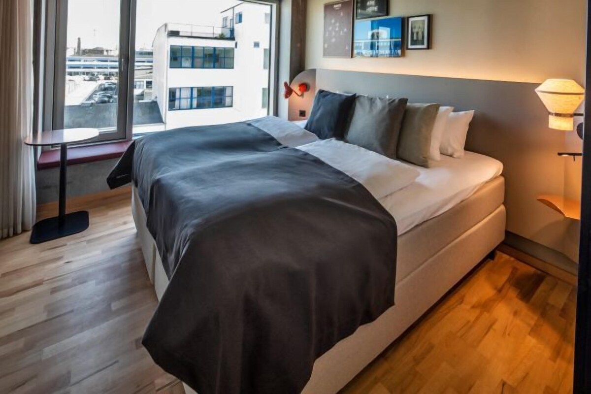Una habitación de hotel moderna con una cama grande, una mesita de noche, una lámpara y vistas a la ciudad a través de amplios ventanales.