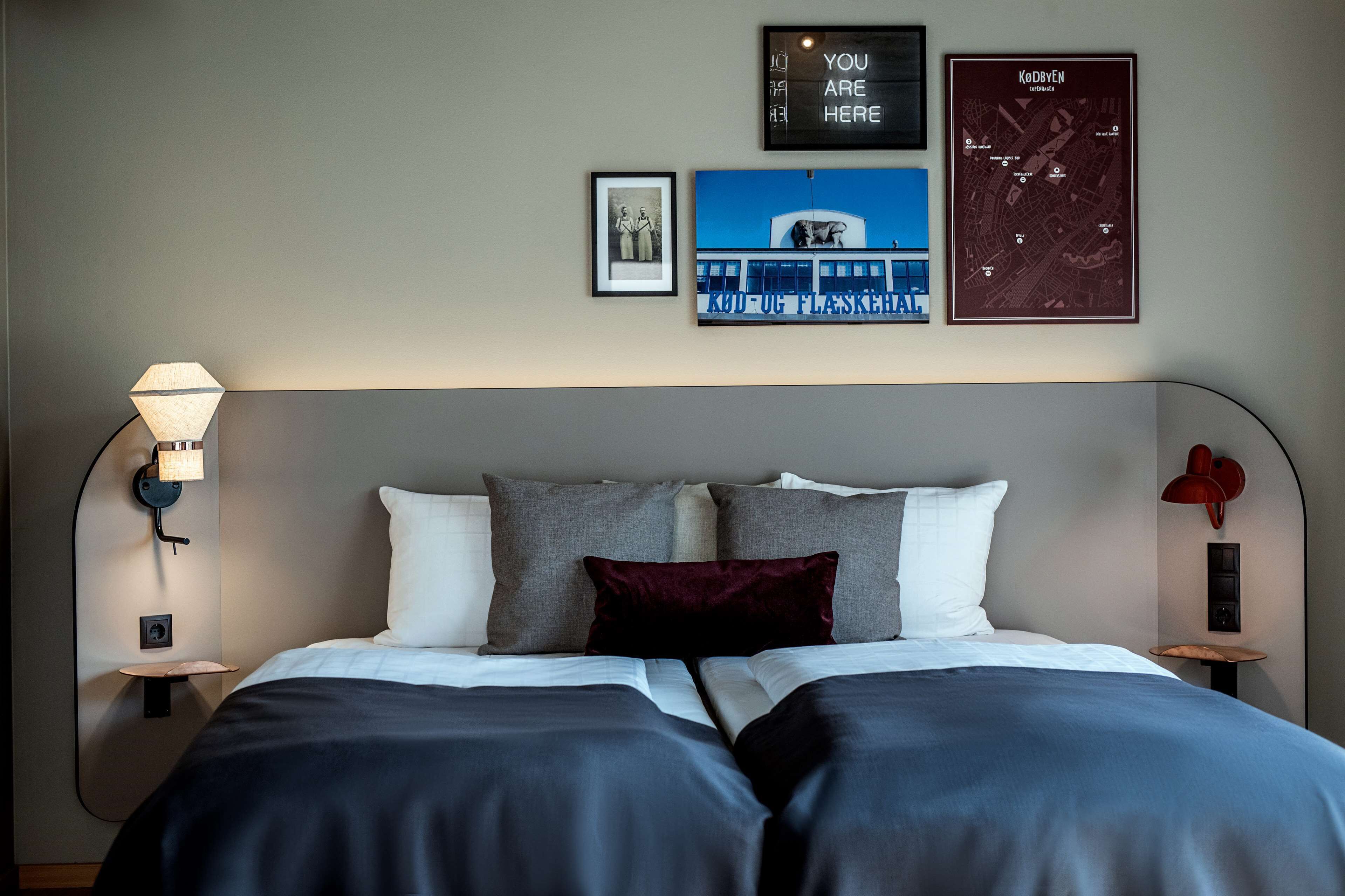 A neatly made bed with a dark blue bedspread, two pillows, and a red accent pillow. There are two wall-mounted lamps on either side of the bed. Above the bed, there are three framed items: a black and white photo, a framed map, and a framed sign with text.