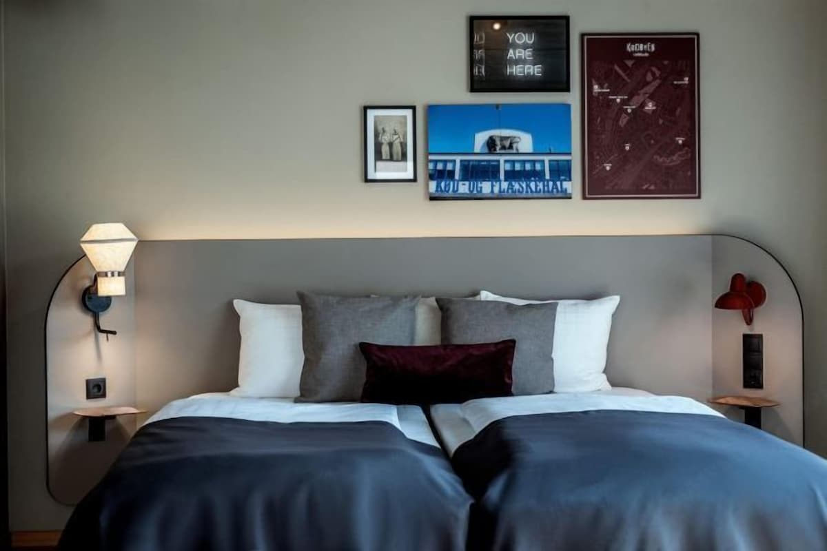 Una cama bien hecha con una colcha azul oscuro y almohadas blancas, una lámpara de pared y cuadros enmarcados que incluyen un mapa y un edificio.