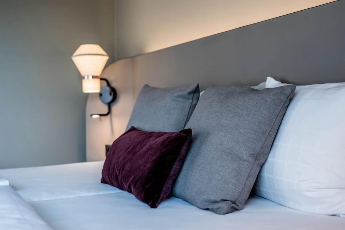 A neatly made bed with white sheets and two pillows, one dark and one light grey, in a room with a grey headboard and a wall-mounted lamp.