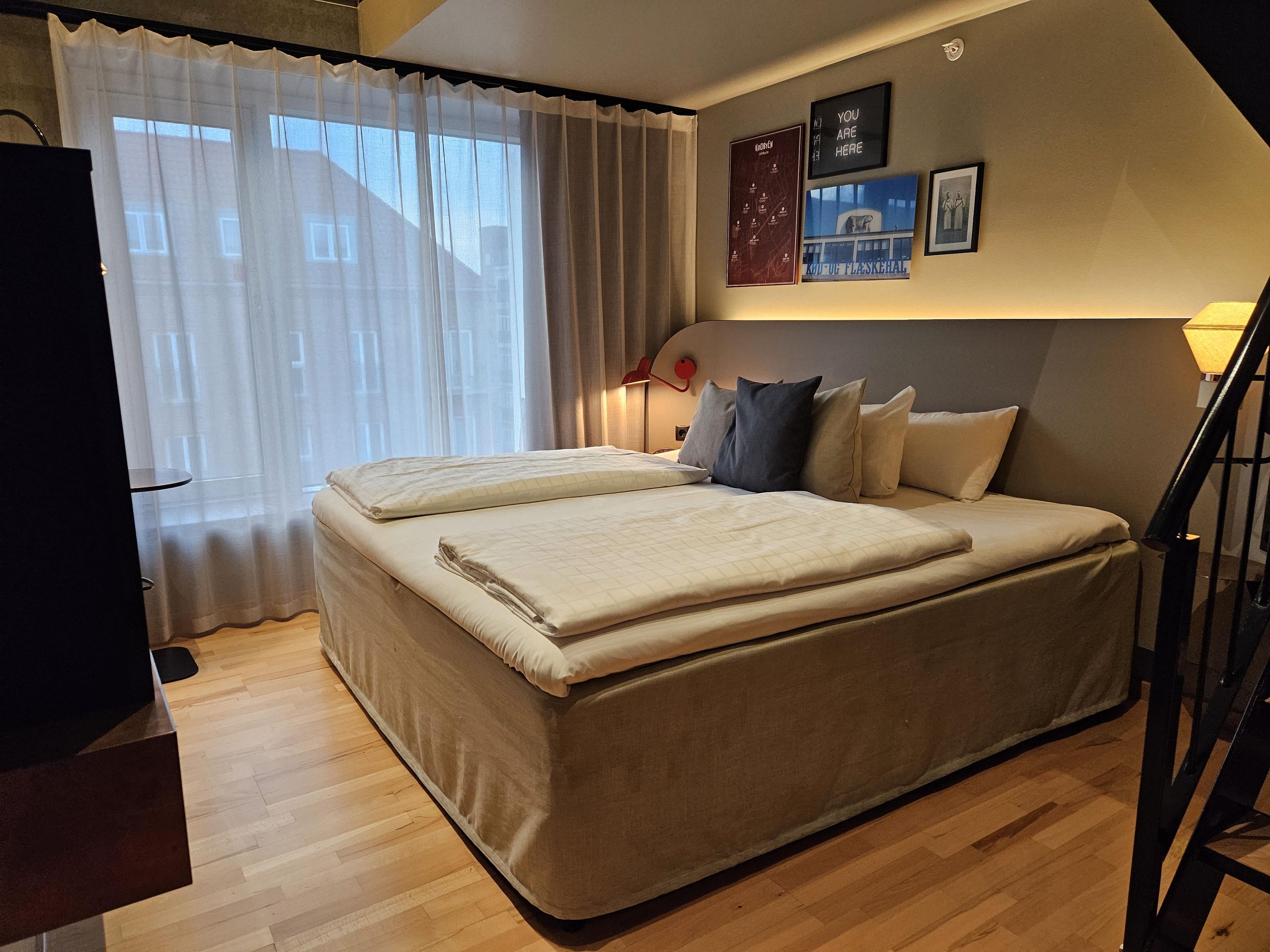 Un dormitorio con una cama grande, una mesita de noche, una lámpara y una ventana con cortinas.