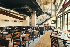 A spacious restaurant with high ceilings, exposed ductwork, and a central staircase. Tables are set with wine glasses and menus.