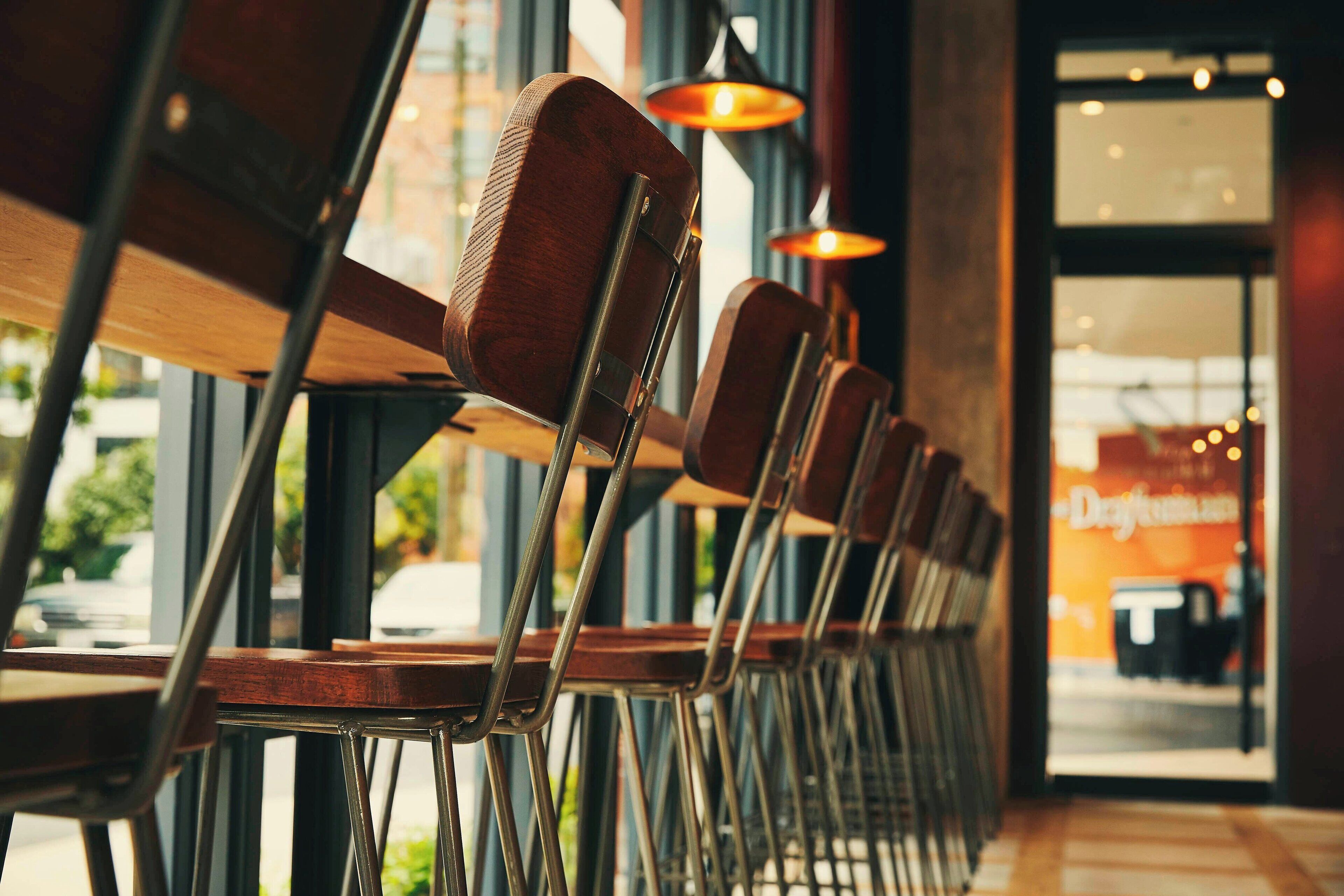 A row of wooden chairs with metal frames, arranged in a restaurant setting with large windows and hanging pendant lights.