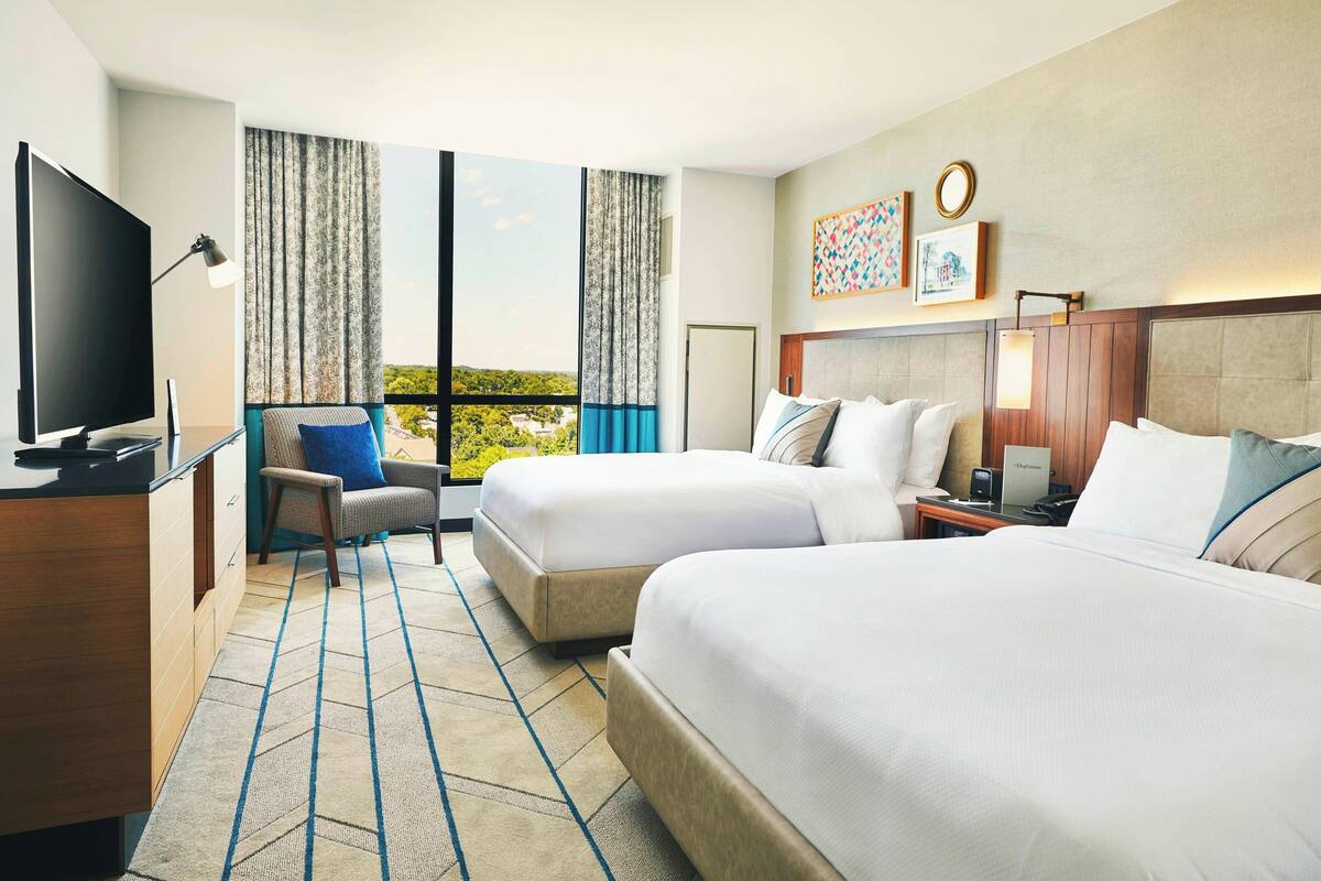 A hotel room with two beds, a TV, a desk, and a view of the outdoors.