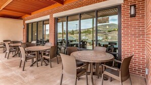 Una terrazza esterna con tavoli rotondi e sedie in vimini, un muro in mattoni e ampie finestre che offrono vista sui dintorni.