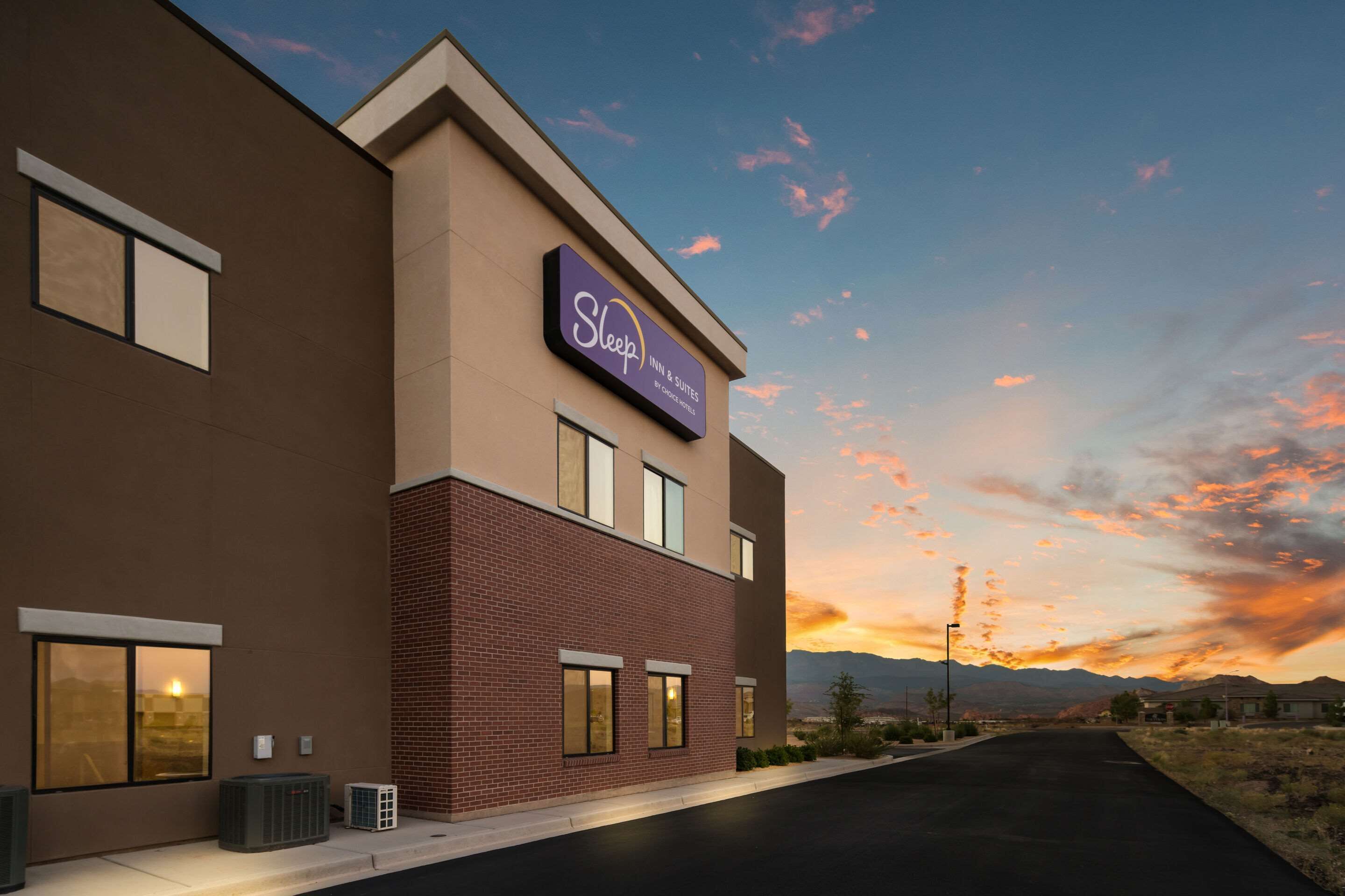 A Sleep Inn & Suites building with a modern design, featuring a prominent sign, large windows, and a view of the surrounding landscape during sunset.