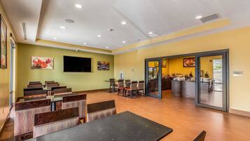A restaurant with wooden tables and chairs, a TV mounted on the wall, and a kitchen area visible through glass doors.
