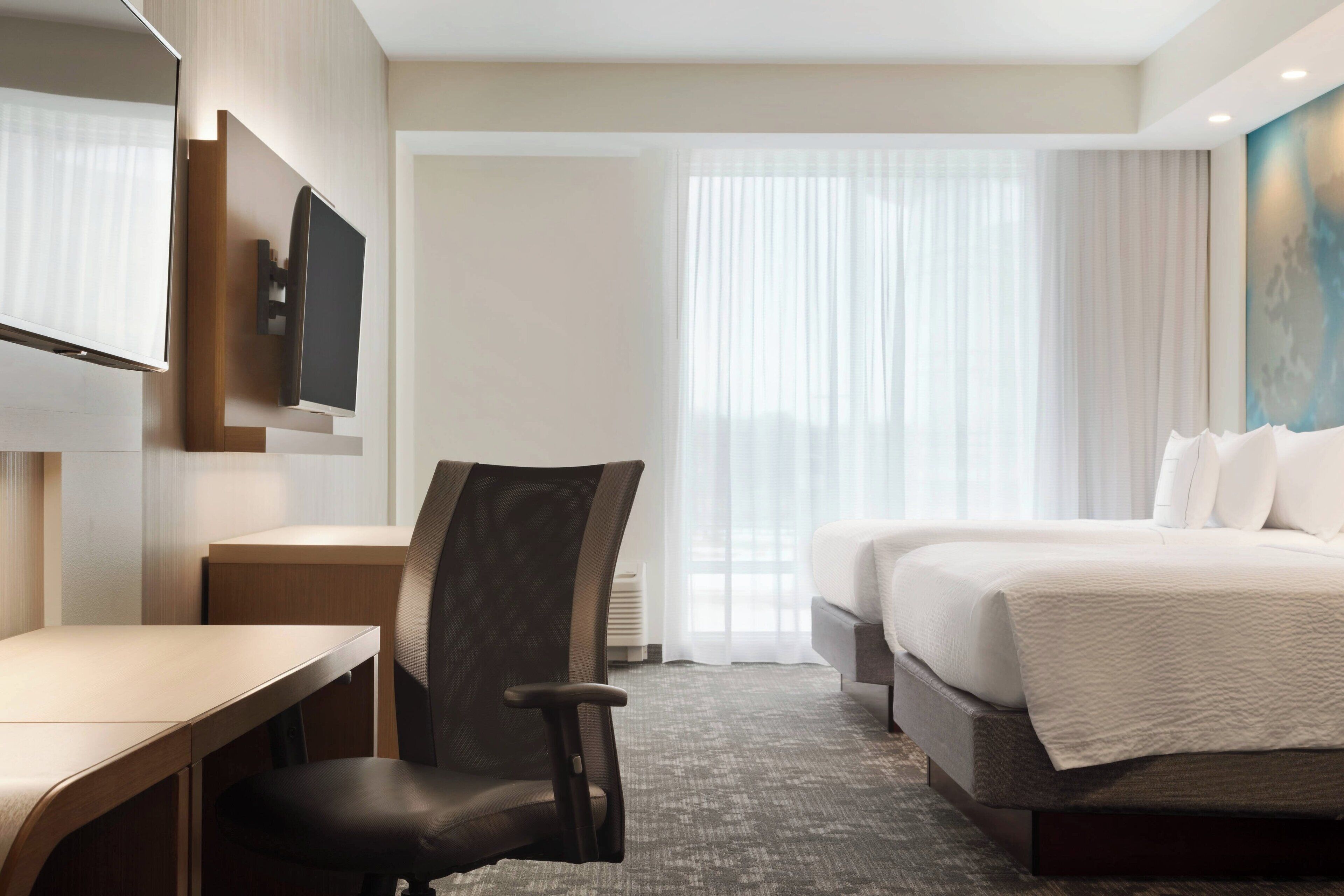 Une chambre d’hôtel comprenant un lit, un bureau, une chaise, un téléviseur fixé au mur et une grande fenêtre avec des rideaux.