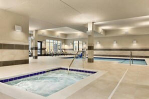 Une grande piscine intérieure dotée de plusieurs couloirs, un espace séparé avec un bain à remous et des chaises longues disposées le long du bord de la piscine.