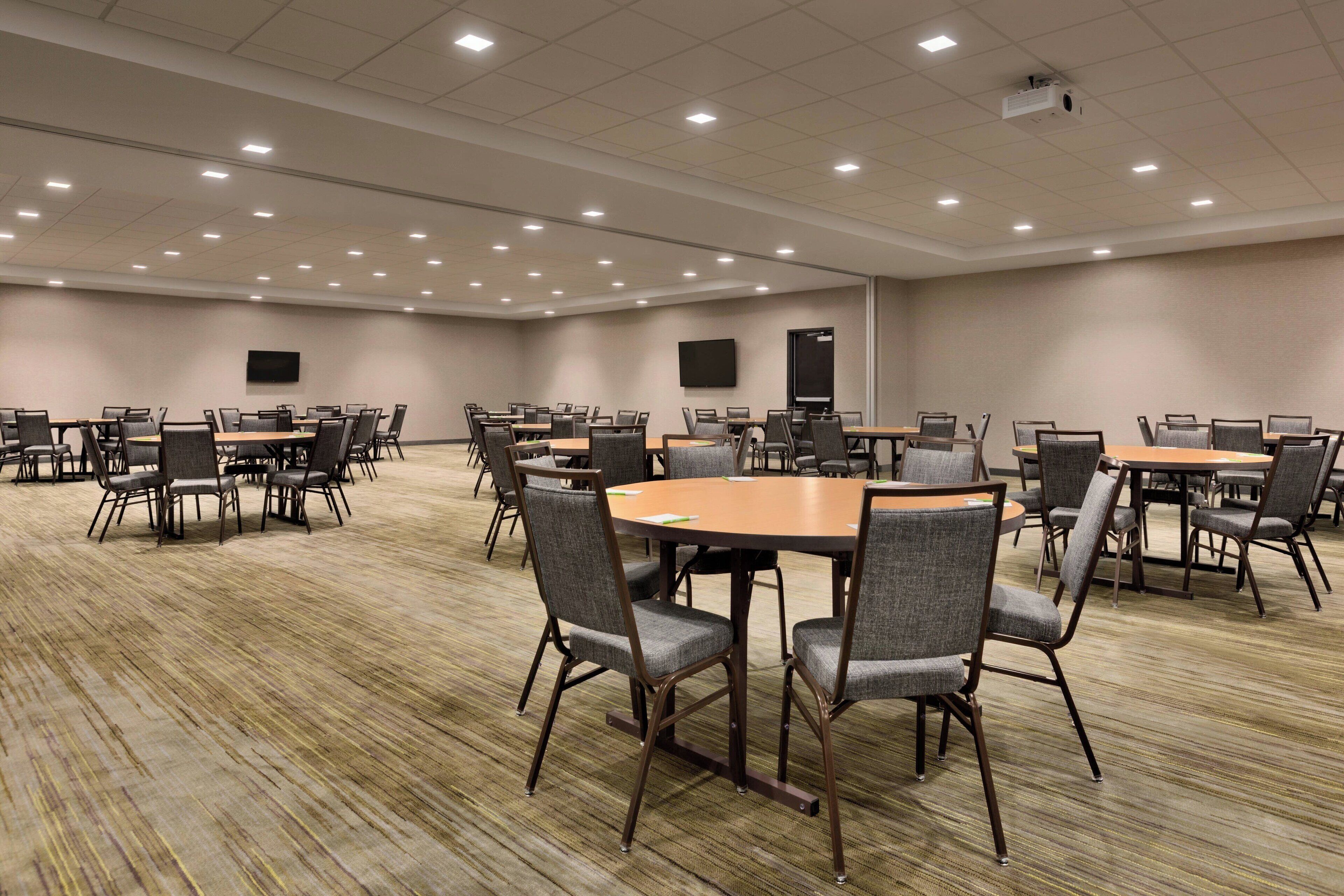 A large conference room with multiple tables and chairs, equipped with ceiling-mounted projectors and flat-screen TVs.