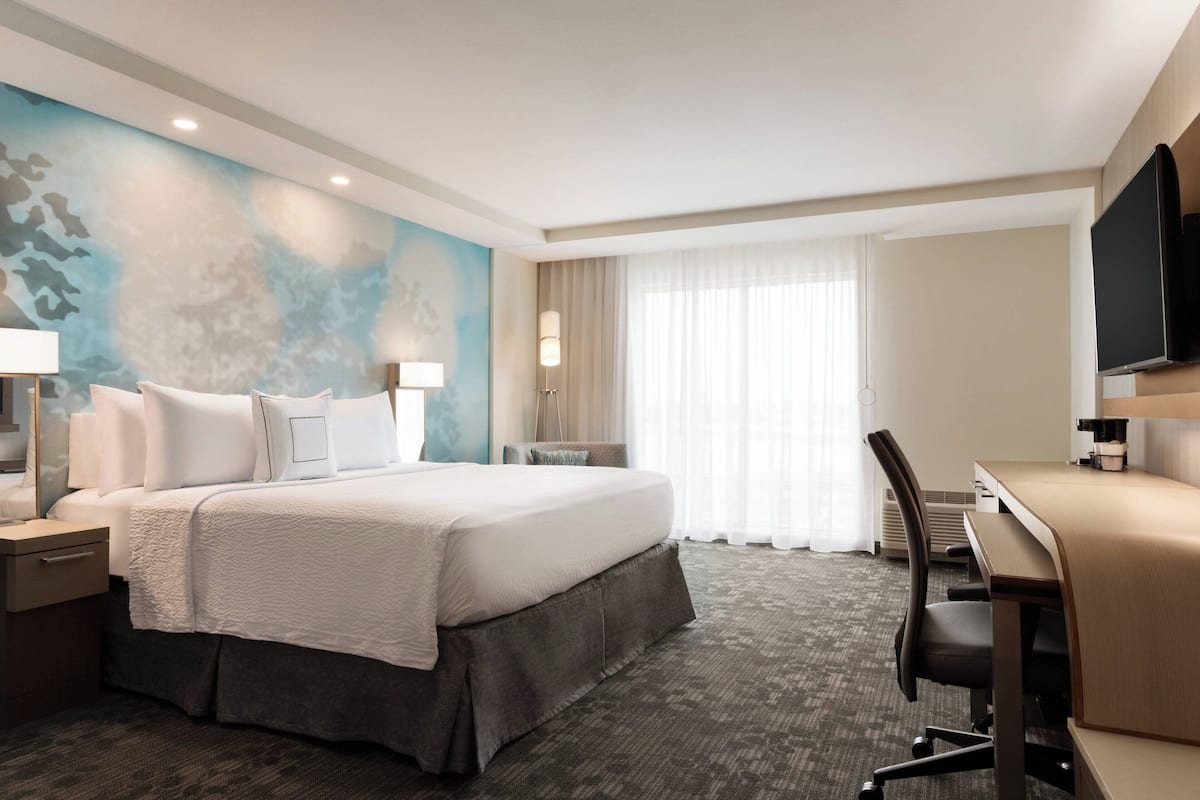 A hotel room with a large bed, a desk, a flat-screen TV, and a large window with sheer curtains.