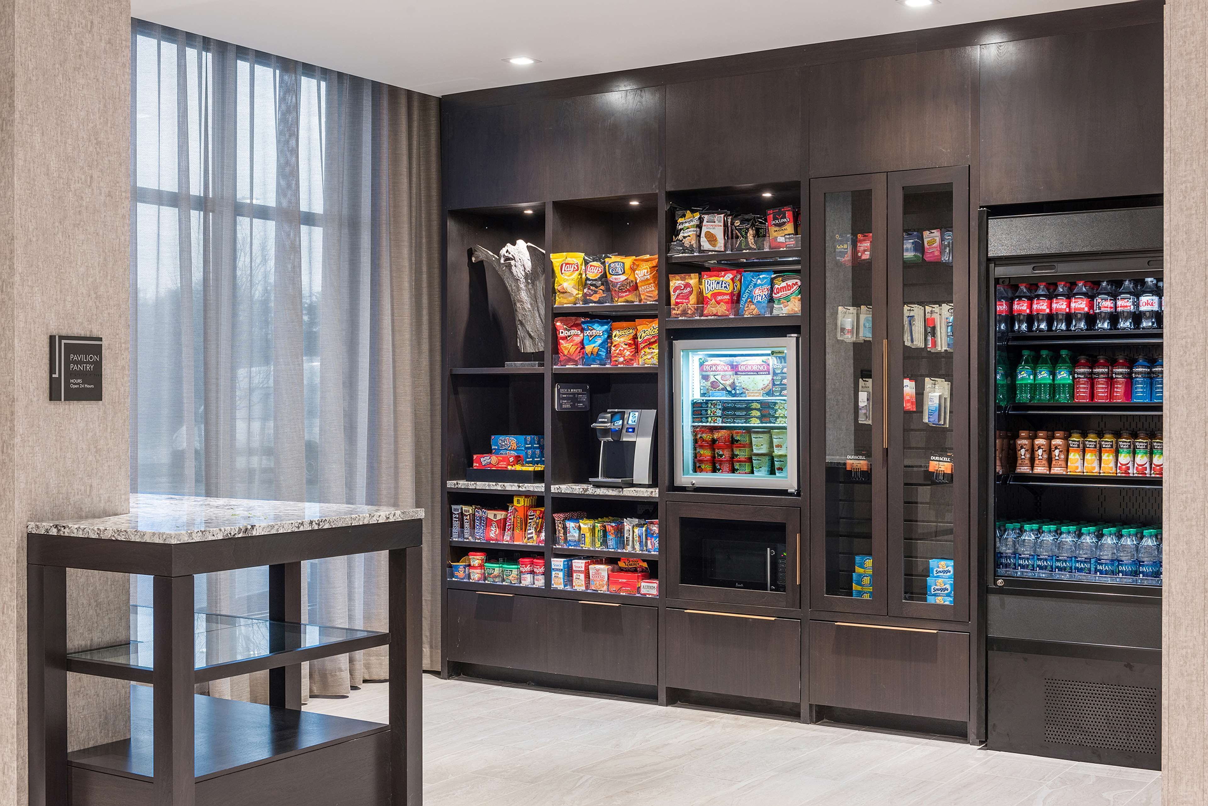A modern hotel pantry with snack and beverage dispensers, a marble-top table, and a view through large windows.