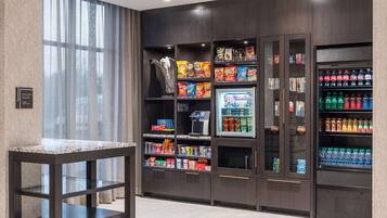 A modern hotel pantry with snack and beverage dispensers, a marble-top table, and a view through large windows.