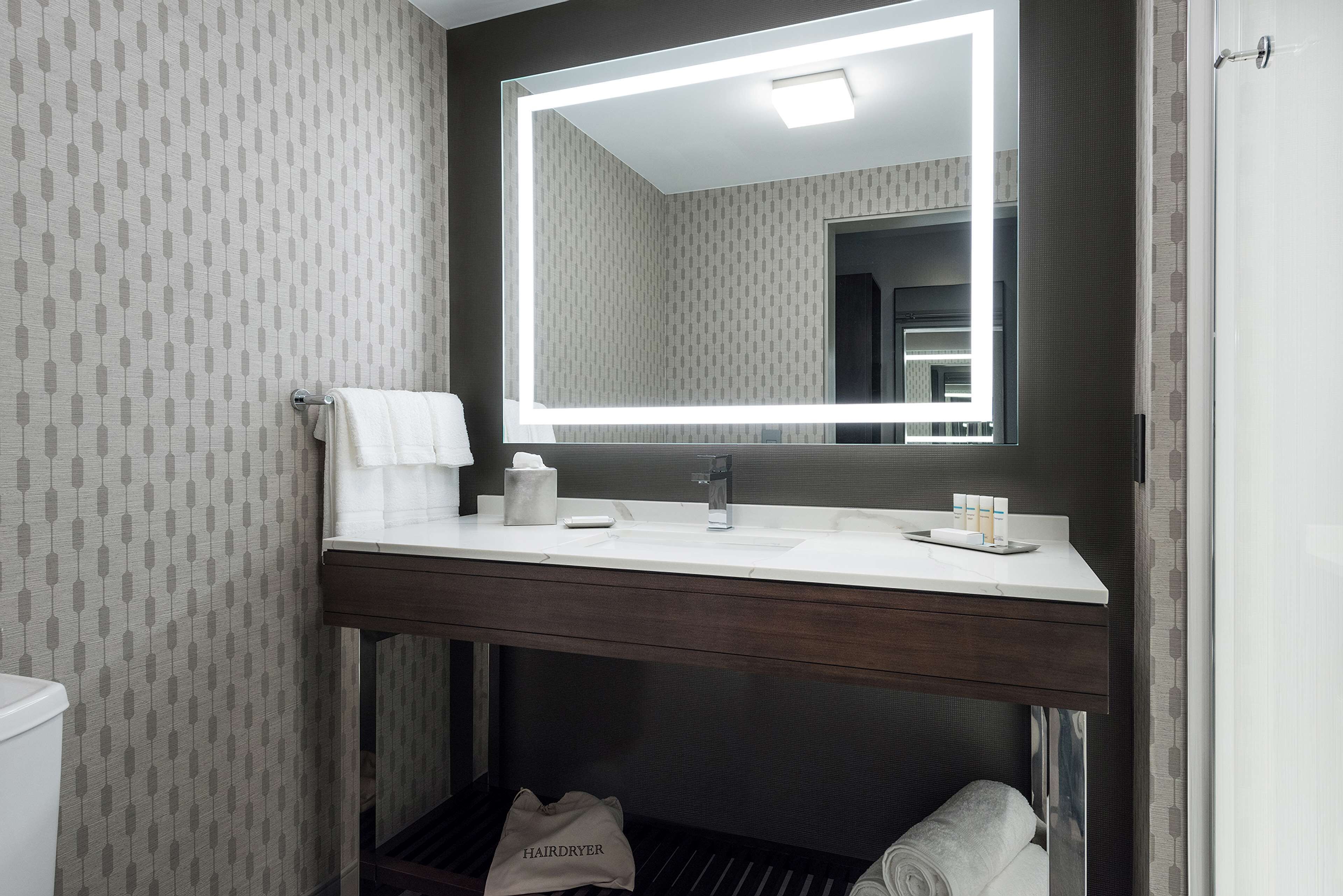 Un baño con tocador, espejo con iluminación integrada, perchero y una bolsa para secador de pelo en el suelo.