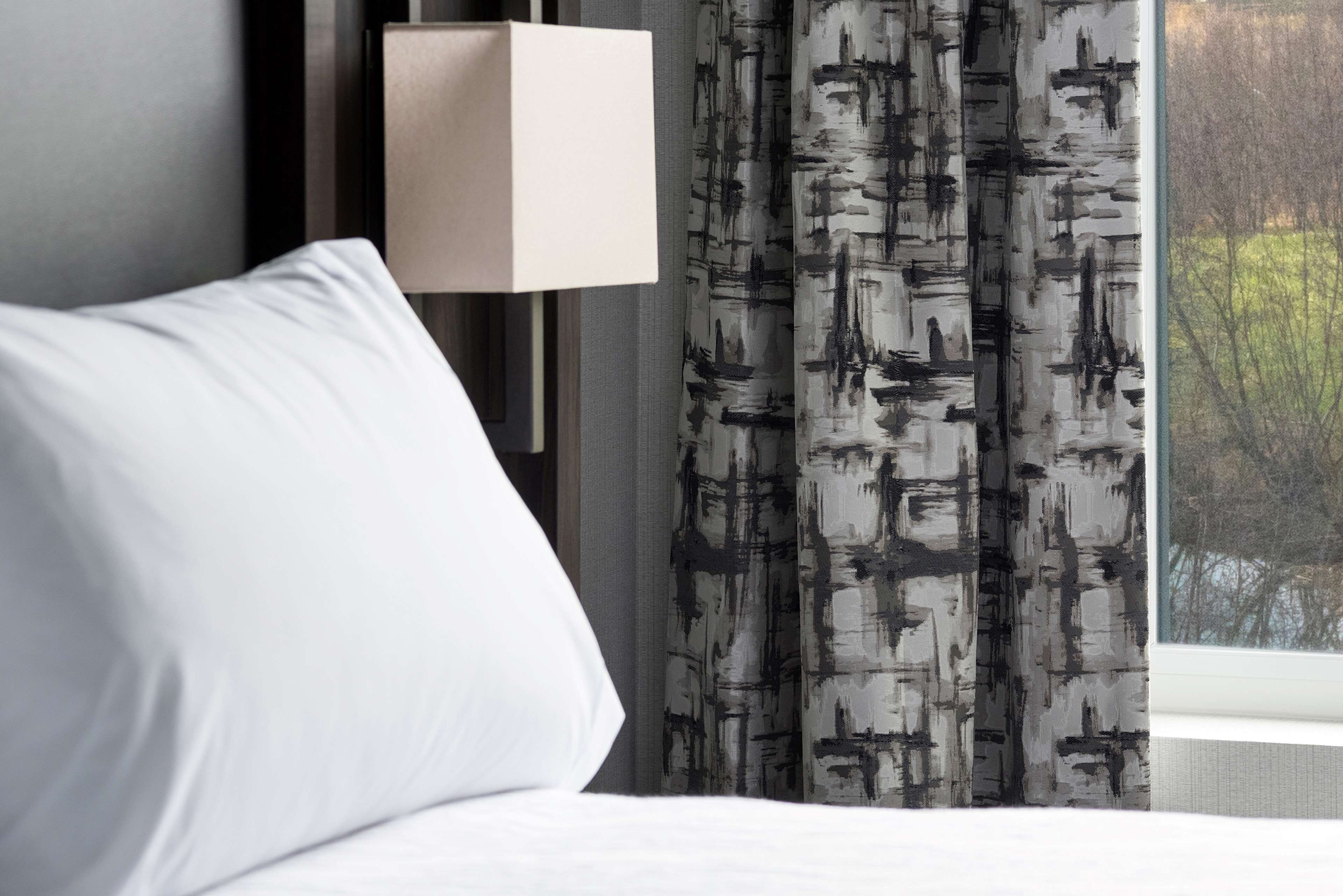 Habitación de hotel con cama blanca, lámpara moderna de pared y ventana con vistas a árboles.
