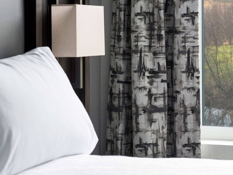 Habitación de hotel con cama blanca, lámpara moderna de pared y ventana con vistas a árboles.