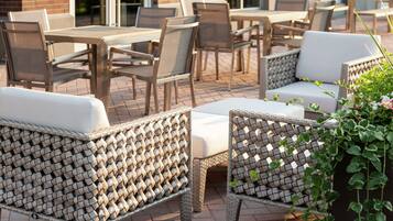 An outdoor seating area with wicker chairs and tables, a brick wall, and a building in the background.
