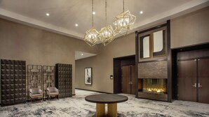 Un hall d’entrée spacieux avec une table ronde, deux fauteuils, un motif géométrique sur le mur, une cheminée et un lustre moderne.
