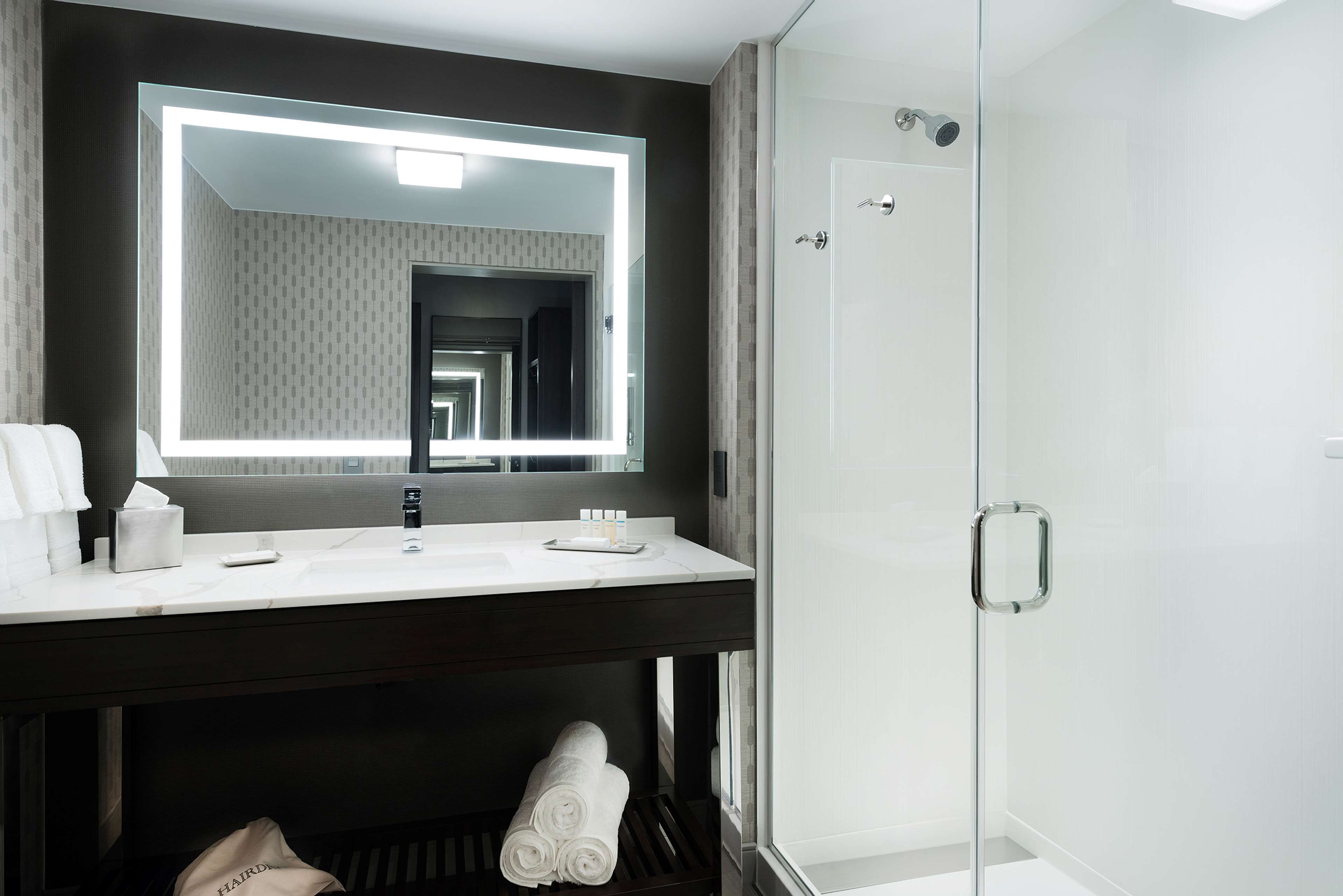 A modern bathroom with a large mirror, a white sink, a glass shower enclosure, and neatly folded towels.