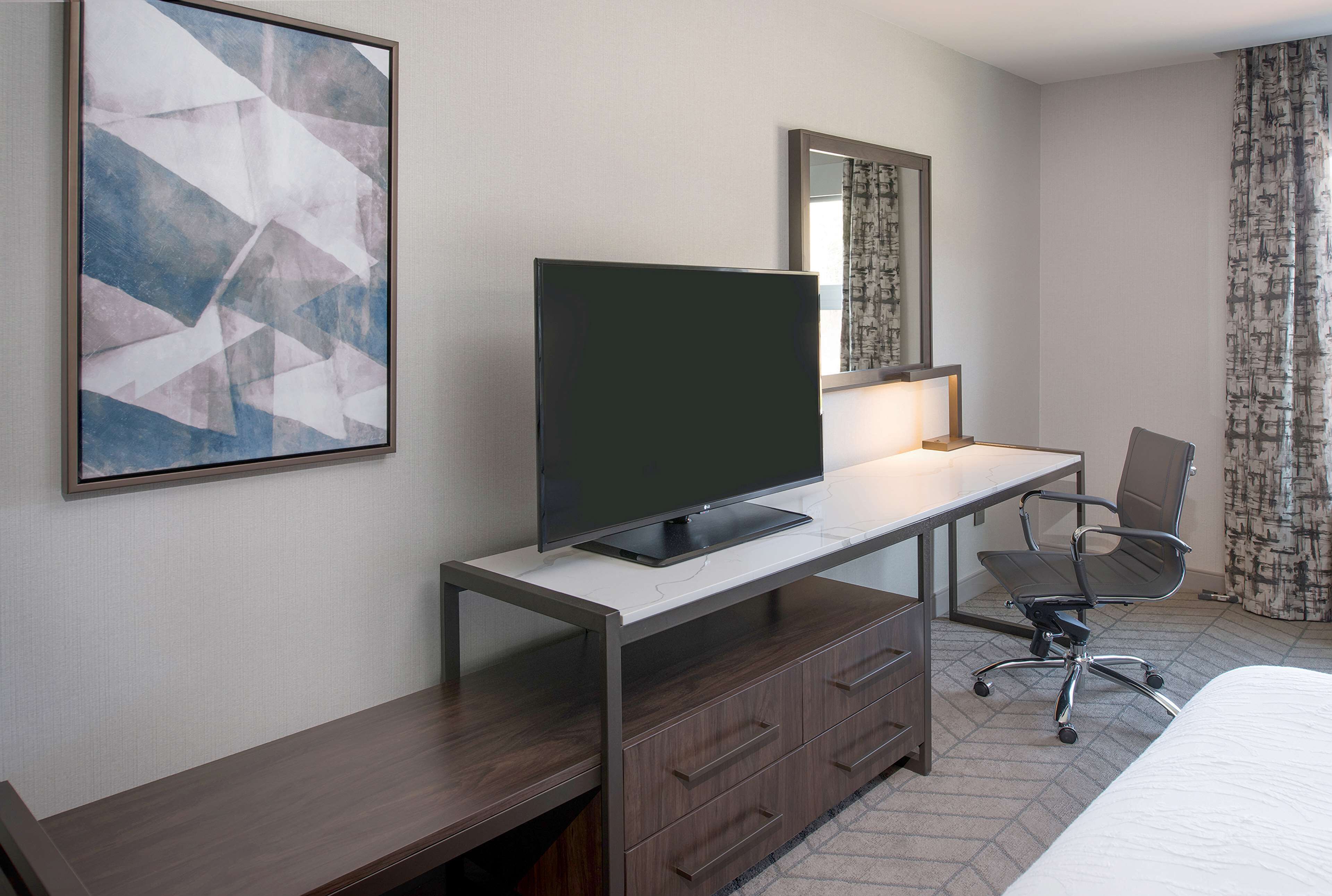 Ein Hotelzimmer mit Bett, Schreibtisch mit Flachbildfernseher, Stuhl, Kommode, Spiegel und einem großen abstrakten Gemälde an der Wand.