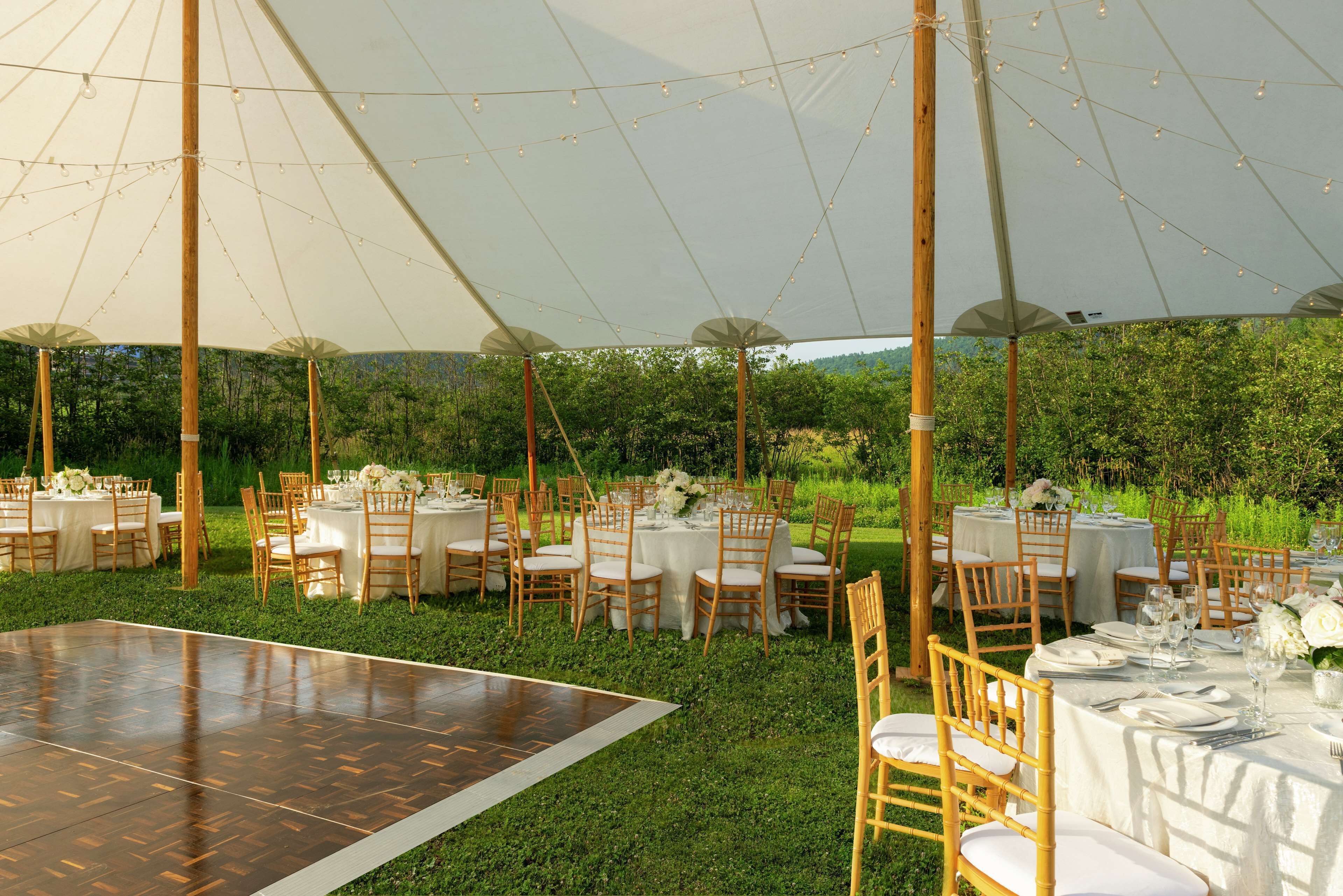 An outdoor wedding venue with round tables, wooden chairs, and a dance floor.