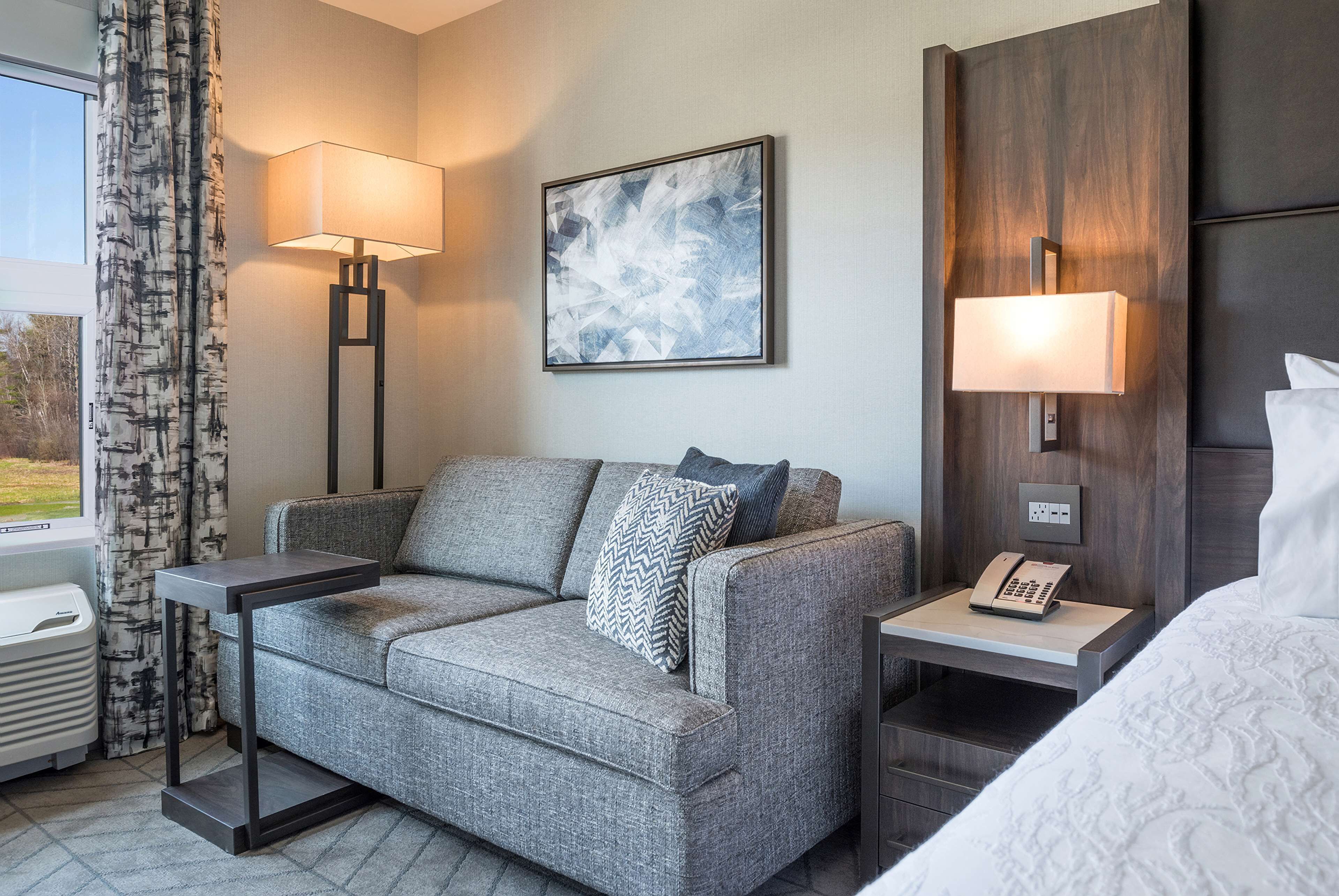 A modern hotel room with a grey sofa, two side tables with lamps, a painting on the wall, and a bed with a white comforter.