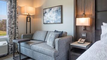 A modern hotel room with a grey sofa, two side tables with lamps, a painting on the wall, and a bed with a white comforter.