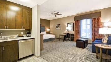 A hotel room with a kitchenette, a bed, a desk, a chair, a sofa, and a ceiling fan.
