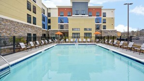 A swimming pool with lounge chairs and umbrellas, surrounded by a stone wall and a multi-story building with balconies.