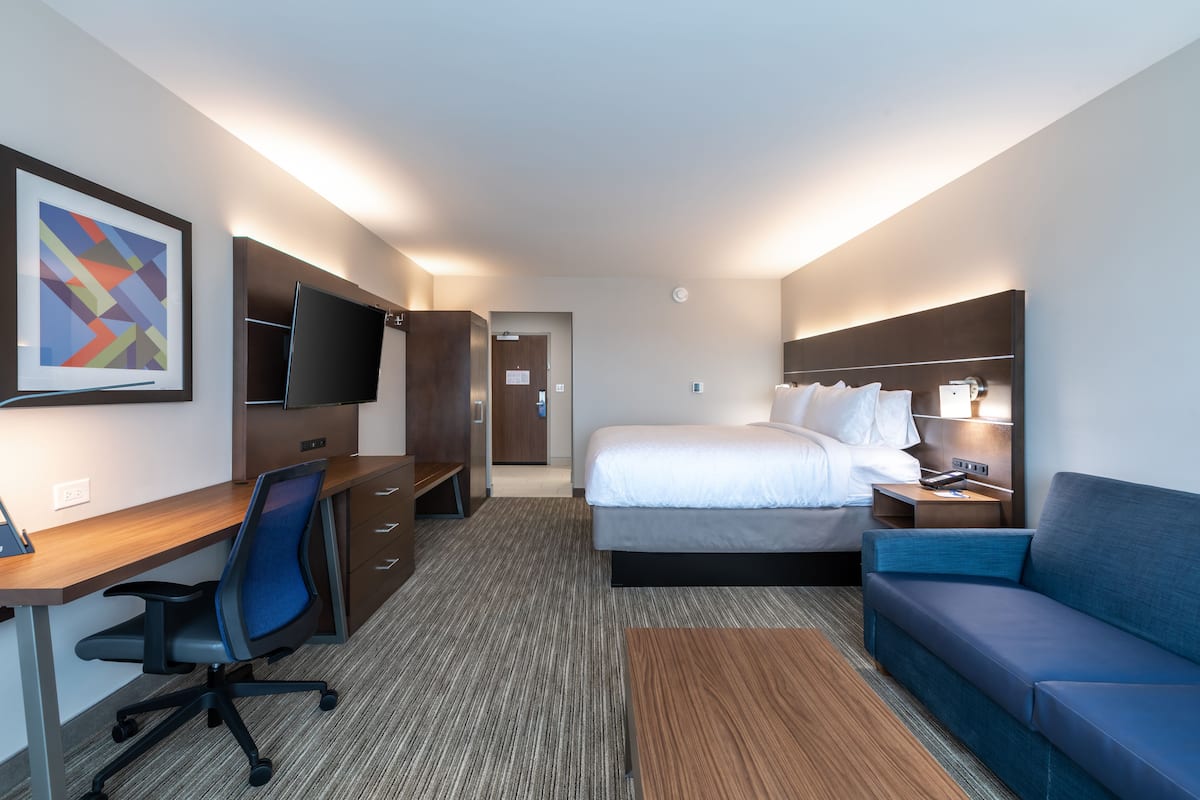 A hotel room with a bed, a desk with a chair, a TV, a sofa, and a framed picture on the wall.