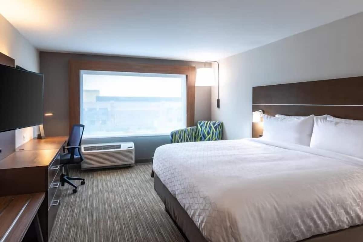 A hotel room with a large bed, a desk with a chair, a television, and a window with a view of buildings.