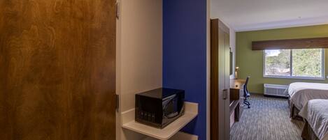 A hotel room with a microwave on a shelf, a bed, a desk with a chair, and a window with a view of greenery.