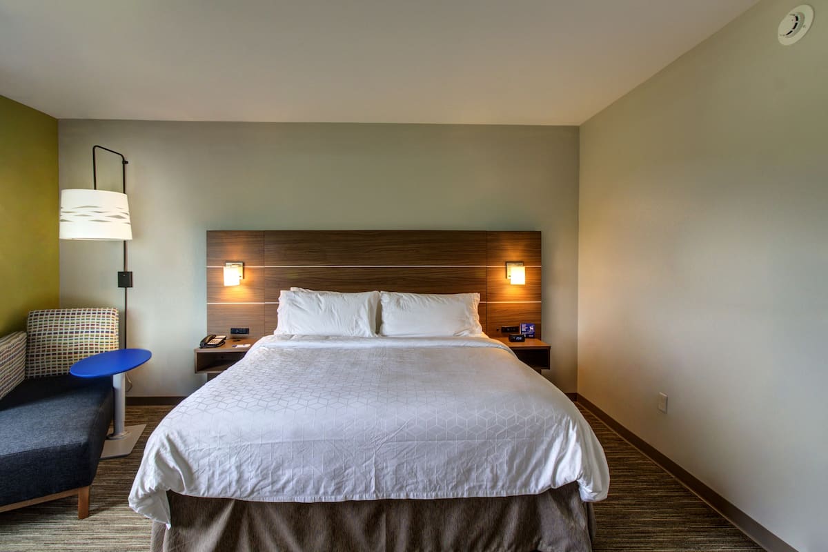 A hotel room with a large bed, a desk with a lamp, a chair, and a bedside table with a phone and a lamp.