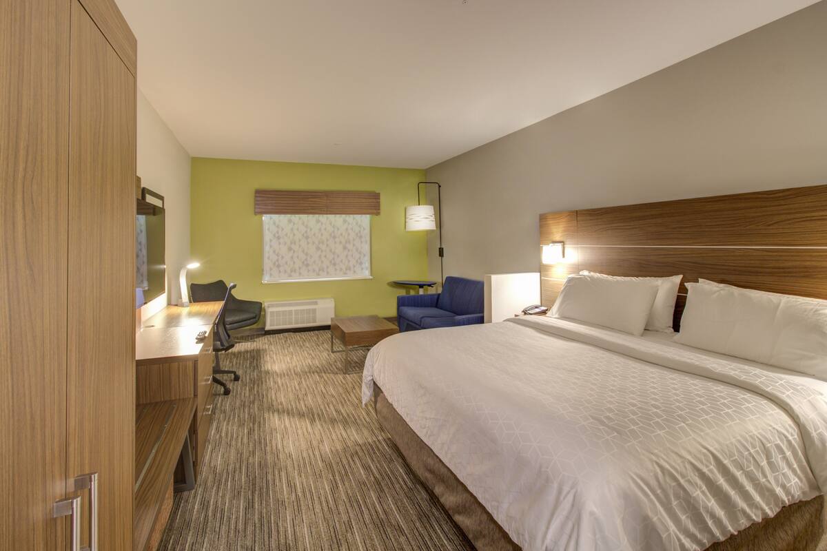 A hotel room with a large bed, a desk with a chair, a small table, and a lamp.